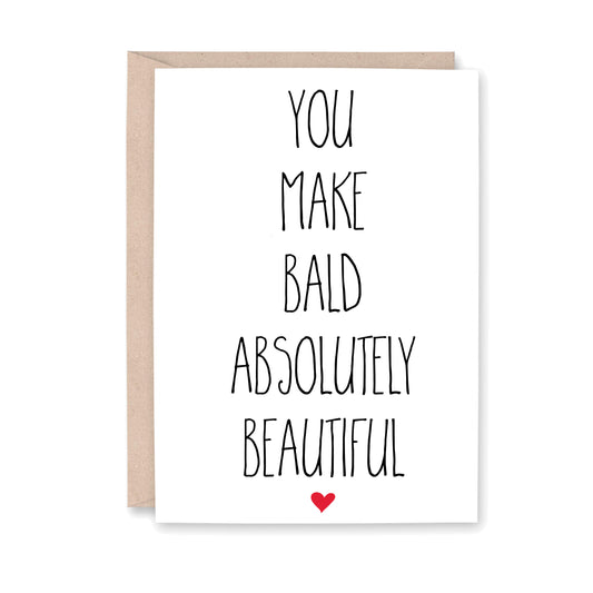 Greeting card that says You make Bald absolutely beautiful, on a white card with a small red heart at the bottom, and a kraft brown envelope in the background